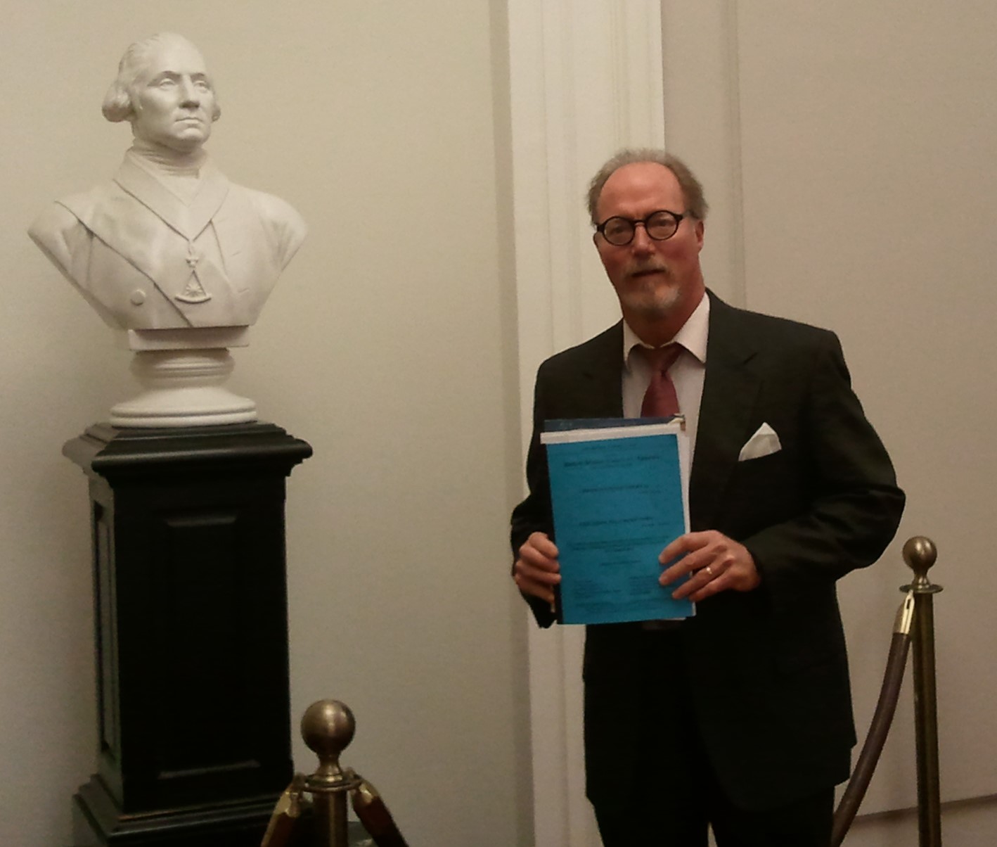 William Auman standing in a suit inside a courthouse hallway holding blue legal documents beside a white sculpted bust on a pedestal, with rope stanchions and neutral walls in the background.